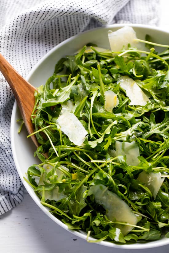 Restaurant Style Arugula Salad is the perfect fresh and light side dish for any meal. This super simple salad is used in restaurants all over the world.ย Arugula salad in a white bowl with a wooden spoon in it.